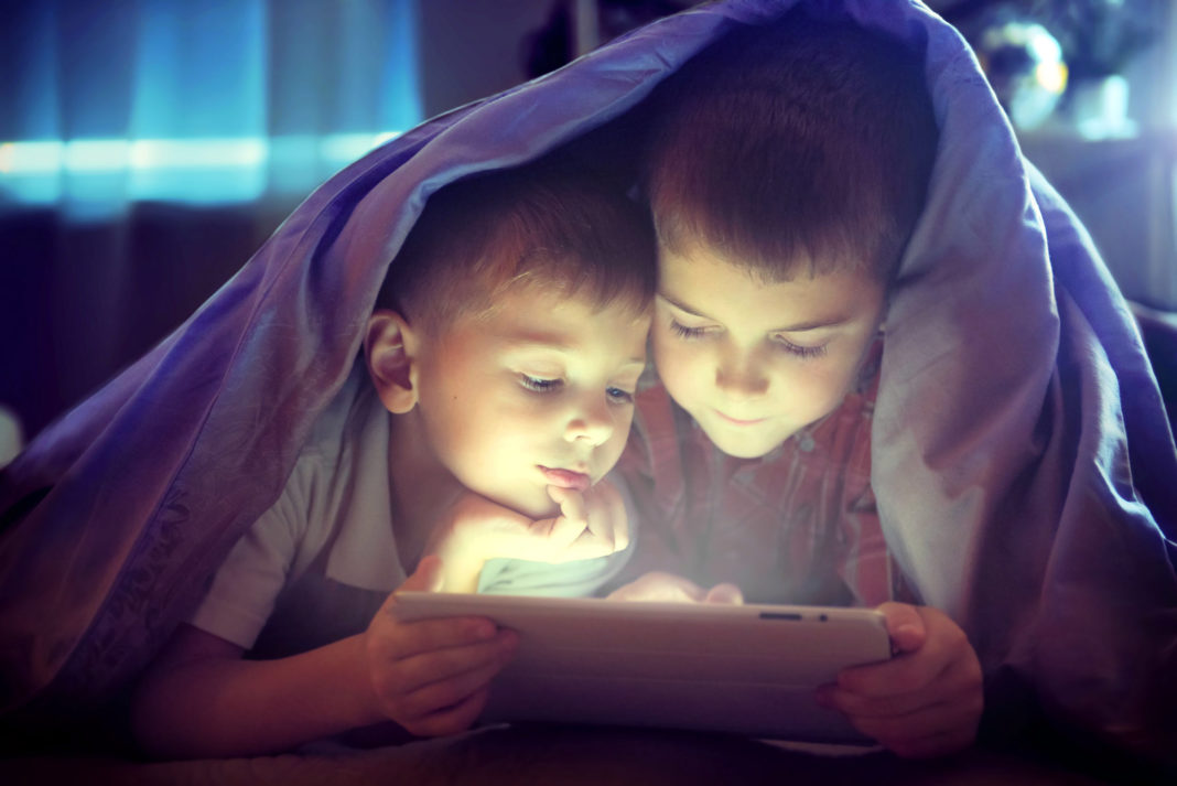 Image of two kids using tablet under blanket at night.